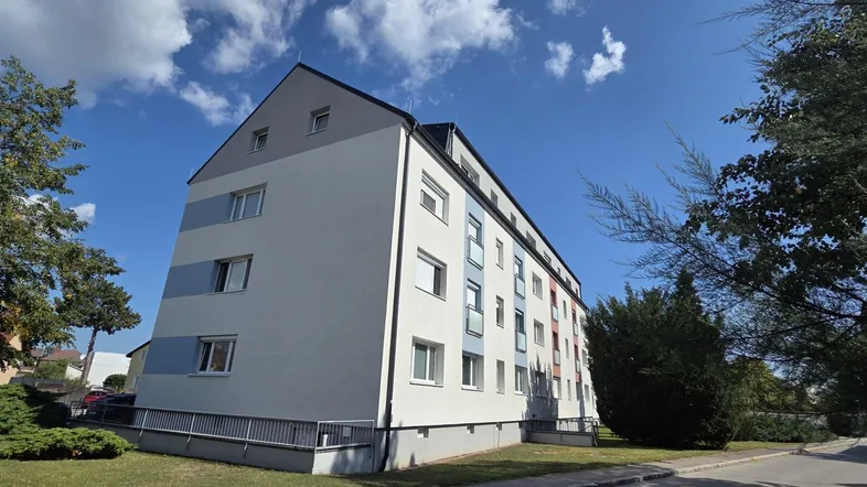 Mehrfamilienhaus mit weißer Fassade und Satteldach, umgeben von Bäumen unter blauem Himmel.