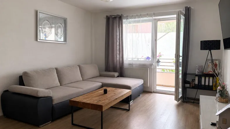 Helles Wohnzimmer mit beigem Ecksofa, Holztisch und Zugang zu einem Balkon.