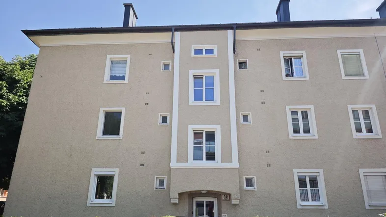 Fassade eines Mehrfamilienhauses mit mehreren Fenstern und gepflegter Hecke im Vordergrund.