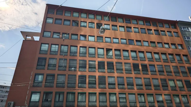 Mehrstöckiges Gebäude mit vielen Fenstern und Geschäften im Erdgeschoss unter blauem Himmel.