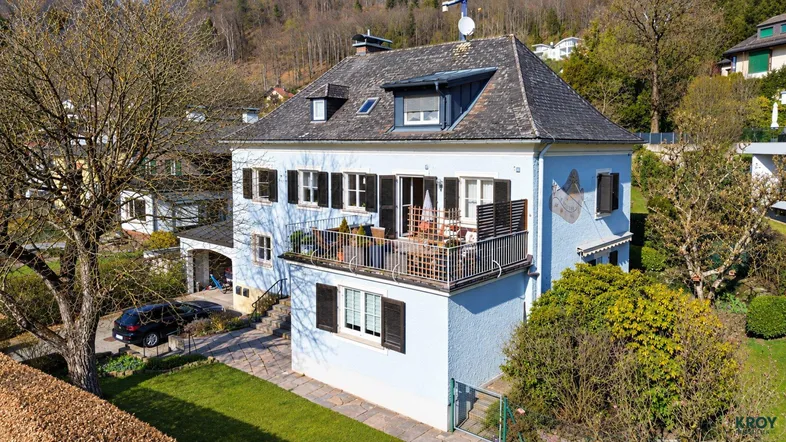 Freistehendes Haus mit hellblauer Fassade, Satteldach und Balkon in grüner Umgebung.