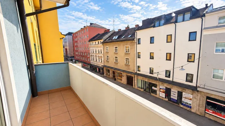 Heller Balkon mit Fliesenboden und Blick auf die belebte Stadtstraße unter blauem Himmel.