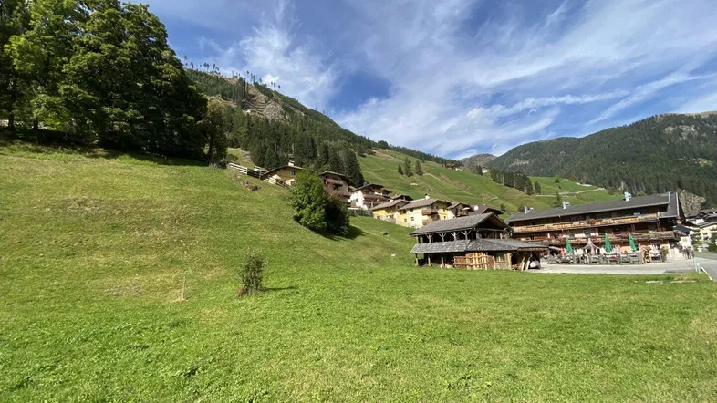 Weitläufige grüne Wiese mit traditionellen Gebäuden und Bergen im Hintergrund unter blauem Himmel.