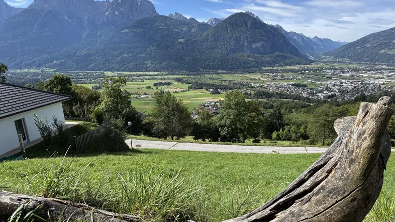Weitläufiger Blick über grüne Hügel, ein Tal mit Siedlung und majestätische Berge im Hintergrund.
