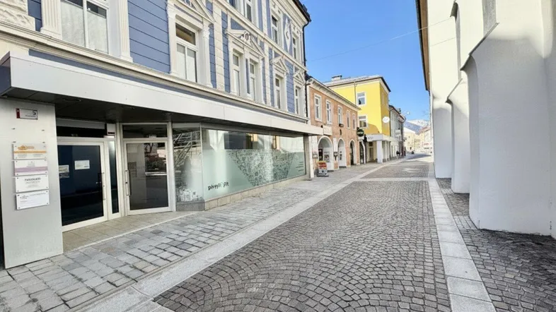 Blick auf die gepflasterte Straße und die historische Fassade des Gebäudes mit Geschäftsräumen im Erdgeschoss.