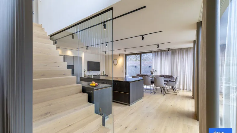 Offener Wohn- und Essbereich mit Holzboden, moderner Treppe und großen Fenstern, die viel Licht hereinlassen.