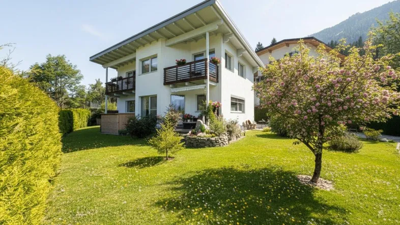 Modernes Wohnhaus mit Balkonen, umgeben von einem grünen Garten und Bergblick unter blauem Himmel.