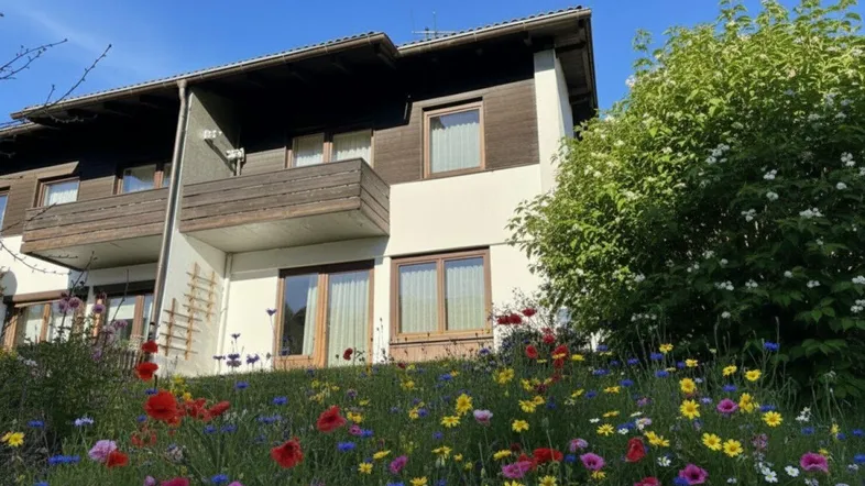 Ansicht des Hauses mit Balkon und einer bunten Blumenwiese im Vordergrund unter blauem Himmel.