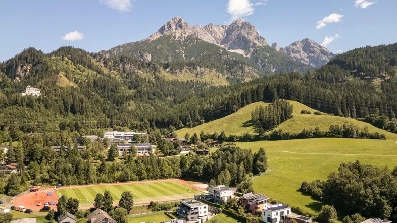 Luftaufnahme einer Wohnsiedlung mit Sportplatz, umgeben von grünen Hügeln und majestätischen Bergen.
