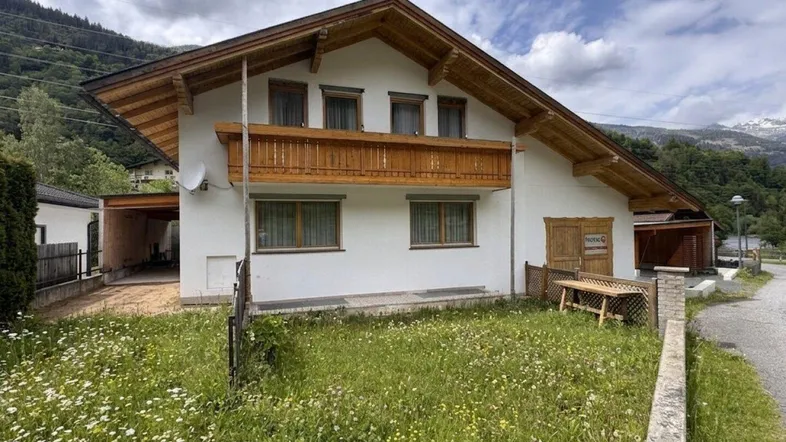 Weißes Haus im alpinen Stil mit Holzbalkon, umgeben von grüner Wiese und Bergen im Hintergrund.