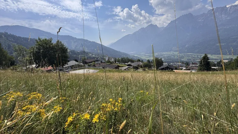 Weitläufige Wiesenlandschaft mit Blick auf ein Dorf und die majestätischen Berge im Hintergrund unter blauem Himmel.
