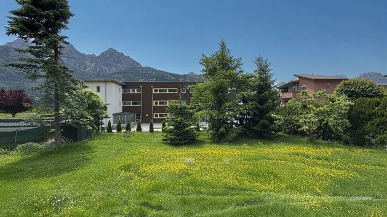 Weitläufige Grünfläche mit blühenden Wiesen und Blick auf die umliegende Berglandschaft und ein modernes Gebäude.