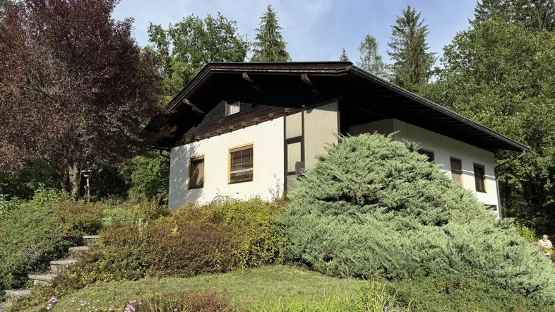 Ländliches Haus mit weißer Fassade und dunklem Dach, umgeben von üppiger Vegetation und Bäumen.