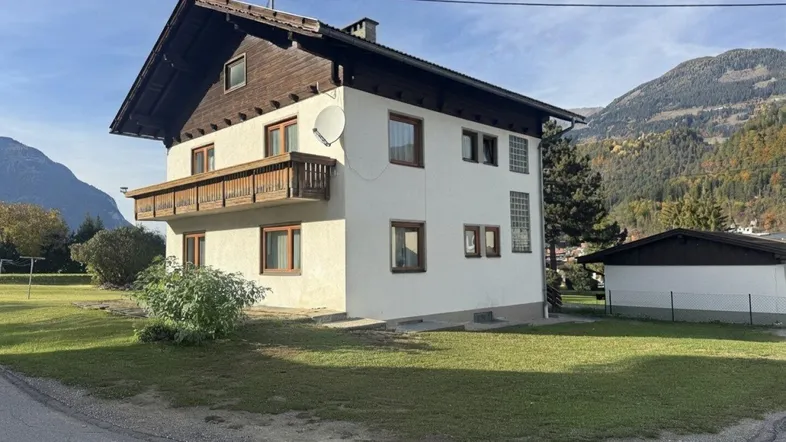 Traditionelles Haus mit Holzbalkon und Satteldach, umgeben von grüner Landschaft und Bergen.