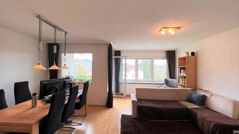 Helles Wohn- und Esszimmer mit Parkettboden, großem Sofa und Zugang zu einem Balkon.