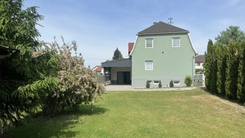 Hinteransicht des Hauses mit großem, gepflegtem Rasen und umgebender Bepflanzung unter blauem Himmel.