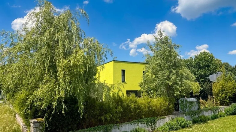 Modernes Haus mit leuchtend gelber Fassade, umgeben von üppigem Grün und Bäumen unter blauem Himmel.