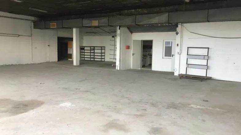 Großer, leerer Raum mit Betonboden, hohen Decken und sichtbaren Deckeninstallationen, ideal als Werkstatt.