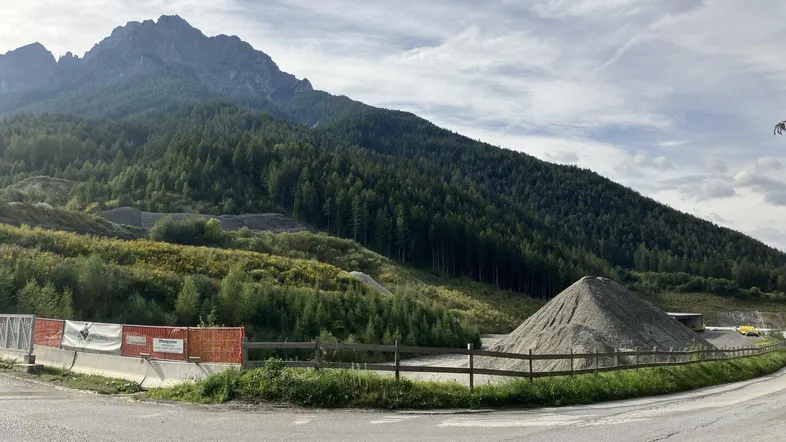 Panoramablick auf eine Berglandschaft mit bewaldeten Hängen und einem Schotterhaufen am Straßenrand unter bewölktem Himmel.