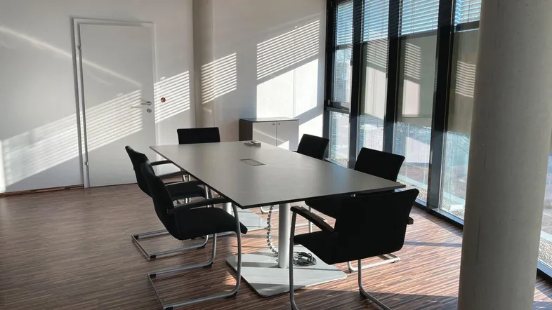 Moderner Konferenzraum mit großem Tisch, bequemen Stühlen und viel natürlichem Licht durch bodentiefe Fenster.