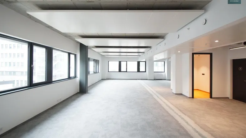 Geräumiges, leeres Büro mit Teppichboden, Sichtbetondecke und großen Fenstern, bereit zur individuellen Gestaltung.