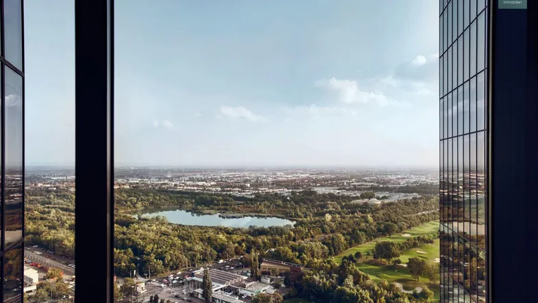 Weitläufiger Ausblick aus einem Hochhausfenster über eine Stadtlandschaft mit See und viel Grün.