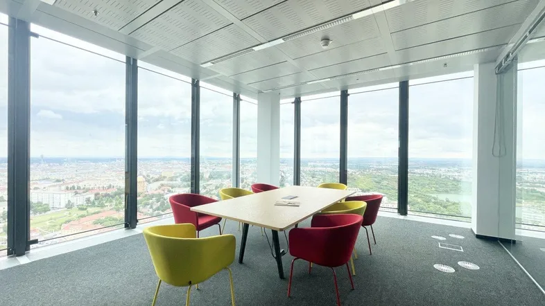 Moderner Besprechungsraum mit großen Fenstern und Panoramablick über die Stadt. Bunte Stühle sorgen für eine lebendige Atmosphäre.