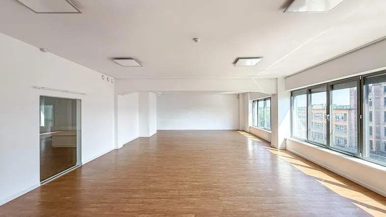 Geräumiger, leerer Büroraum mit großen Fenstern, hellem Holzboden und modernen Deckenleuchten.