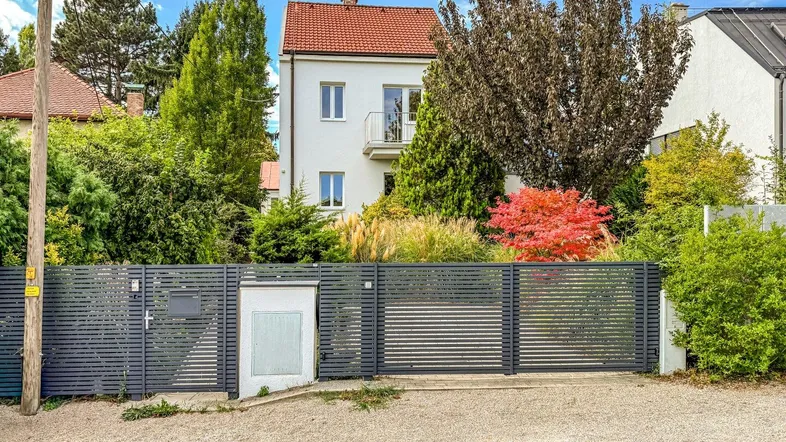 Weißes Wohnhaus mit rotem Dach und modernem Zaun, umgeben von üppiger Vegetation.