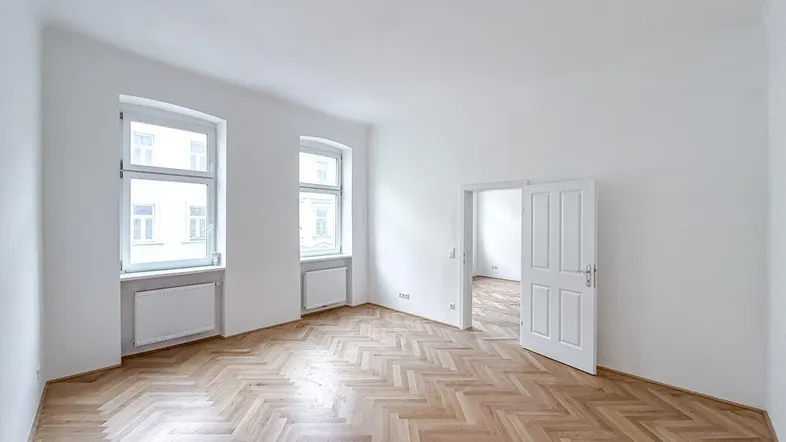 Heller Raum mit Fischgrätparkett und zwei großen Fenstern, die viel Tageslicht hereinlassen.