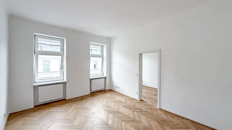 Heller Raum mit Fischgrätparkettboden und weißen Wänden, bietet viel natürliches Licht durch zwei Fenster.