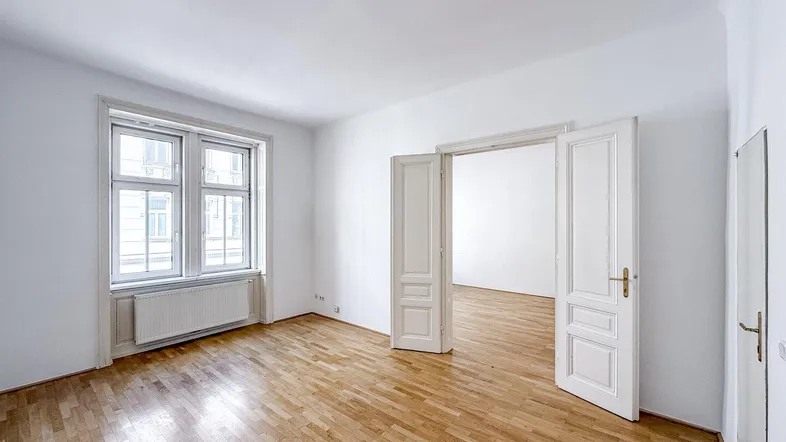 Heller Wohnraum mit Holzboden und großen Fenstern, der zu einem weiteren Zimmer führt.