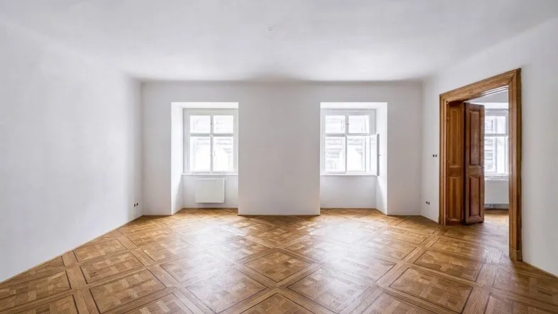 Geräumiges Wohnzimmer mit hellem Parkettboden und zwei großen Fenstern, die viel Tageslicht hereinlassen.