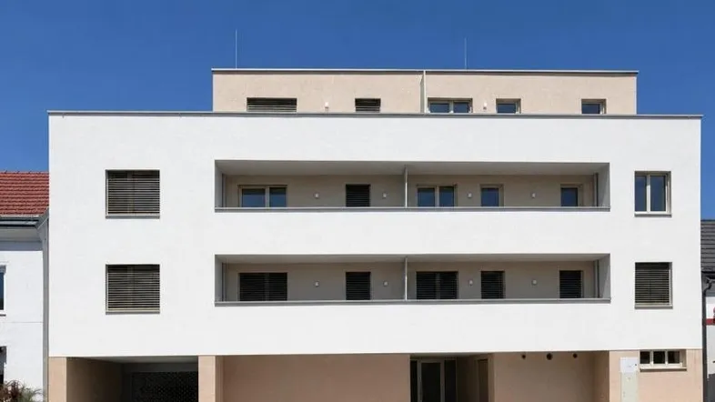 Modernes Mehrfamilienhaus mit weißer Fassade, integrierten Balkonen und Tiefgaragenzufahrt unter blauem Himmel.