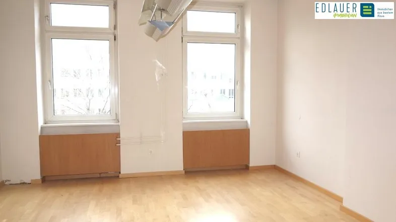 Heller, leerer Raum mit zwei großen Fenstern, Holzfußboden und Heizkörpern, bereit zur Renovierung.