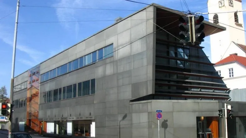 Modernes Geschäftsgebäude mit markanter Betonfassade und großen Fenstern an einer städtischen Kreuzung.