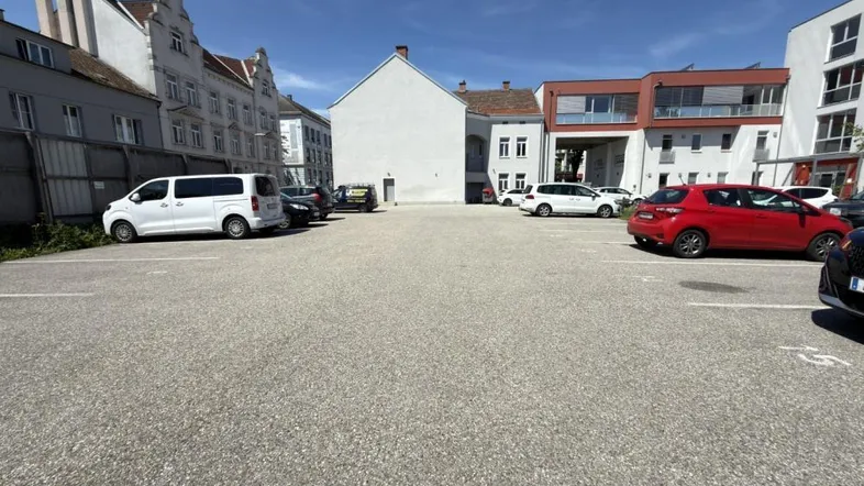 Großer asphaltierter Parkplatz mit umliegenden Gebäuden und geparkten Fahrzeugen unter blauem Himmel.