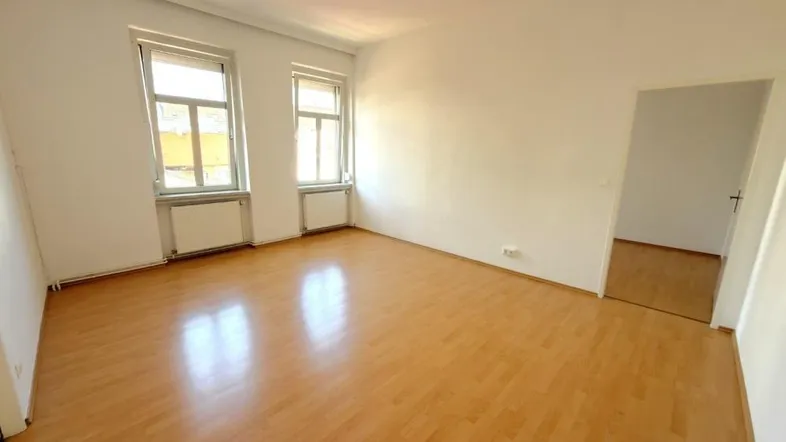 Helles, leeres Zimmer mit Laminatboden und zwei Fenstern, bereit zur individuellen Gestaltung.