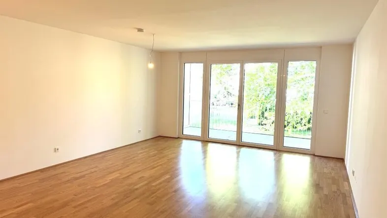 Heller, leerer Raum mit Parkettboden und großen Fenstern mit Blick ins Grüne.