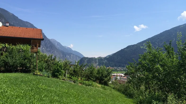 Haus mit rotem Ziegeldach auf einem grünen Hügel mit Obstbäumen und weitem Bergblick.