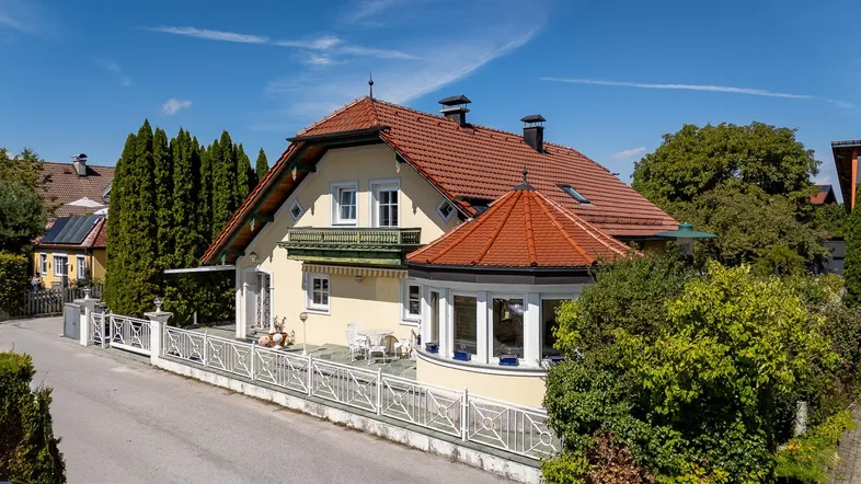 Charmantes Haus mit rotem Ziegeldach, sonniger Terrasse und gepflegtem Gartenbereich unter blauem Himmel.