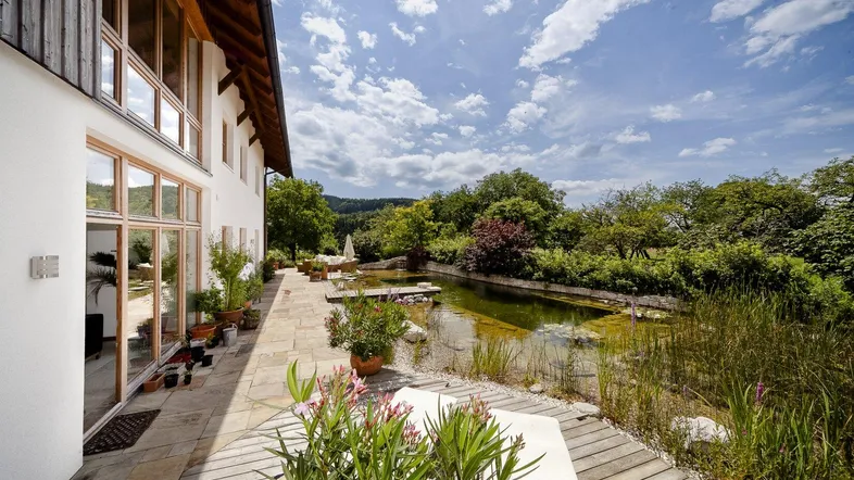 Große Terrasse am Haus mit Naturteich und üppiger Bepflanzung unter blauem Himmel.