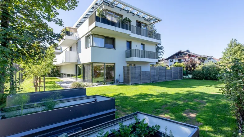 Modernes Mehrfamilienhaus mit gepflegtem Gartenbereich und Hochbeeten unter blauem Himmel.