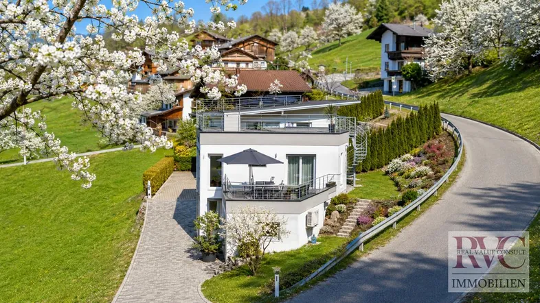 Modernes Haus mit mehreren Terrassen und gepflegter Gartenanlage in einer grünen Hügellandschaft.