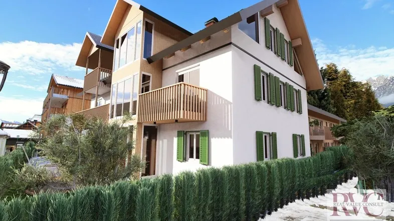 Modernes Gebäude mit Holzverkleidung, grünen Fensterläden und Balkonen vor winterlicher Bergkulisse.