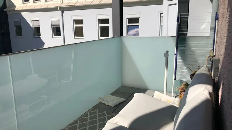 Gemütlicher Balkon mit Glasbrüstung und bequemen Sitzgelegenheiten, bietet einen Blick auf die umliegenden Gebäude.