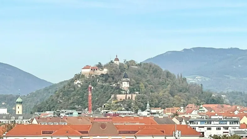 Panoramablick über die Stadt mit dem Schlossberg und umliegenden Hügeln unter klarem Himmel.