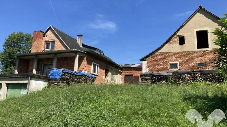 Zwei Backsteingebäude in ländlicher Umgebung mit Holzstapeln und grüner Wiese unter blauem Himmel.