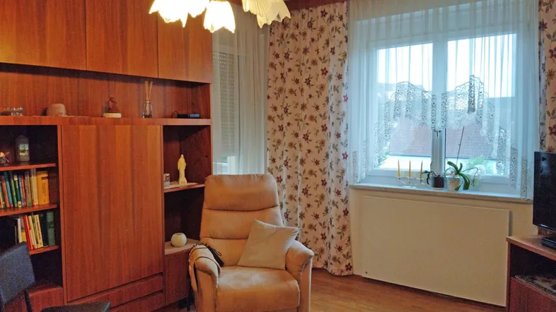 Gemütliches Wohnzimmer mit Holzmöbeln, bequemen Sessel und Fenster mit floralen Vorhängen.