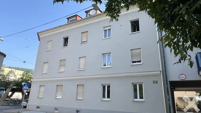 Graue Fassade eines Mehrfamilienhauses mit mehreren Fenstern und Rollläden unter blauem Himmel.